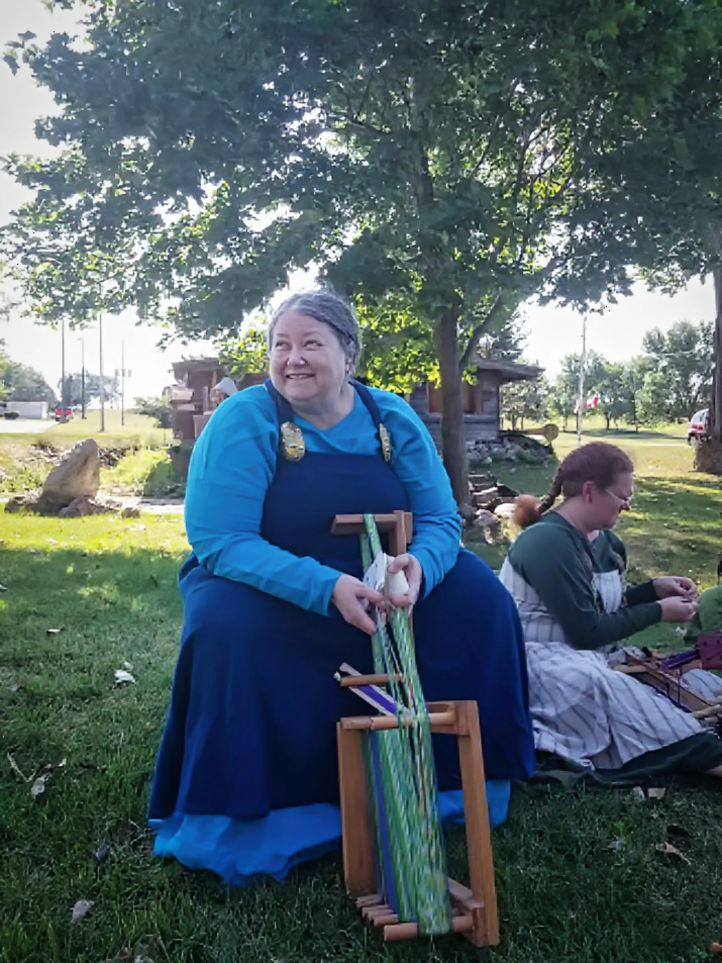 Viking-era costumed woman seated outdoors beside weaving tools