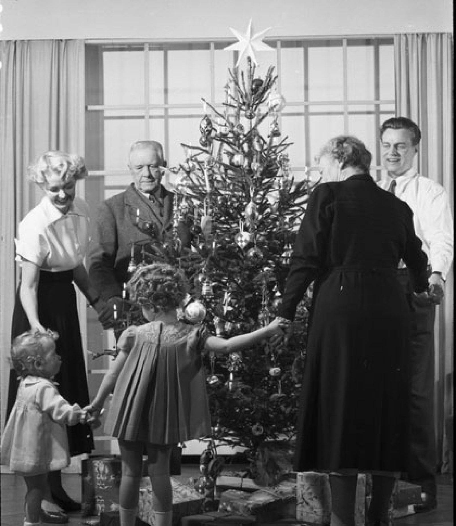 Black and white photograph of a family holding hands around a decorated Christmas tree.